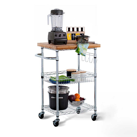 A three-tiered chrome rolling kitchen cart features a wooden butcher-block top holding a blender, small oil bottles, a bowl, and utensils. The lower wire shelves store canister jars, folded dish towels, a large stockpot, and a bowl filled with fresh fruit.
