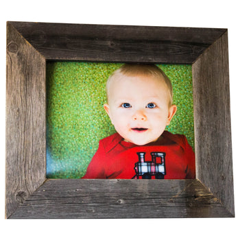 A framed photo shows a baby with fair skin and blue eyes looking directly at the viewer. The baby is wearing a red long-sleeved shirt with a black and white train design on the front and is positioned against a blurred green background.