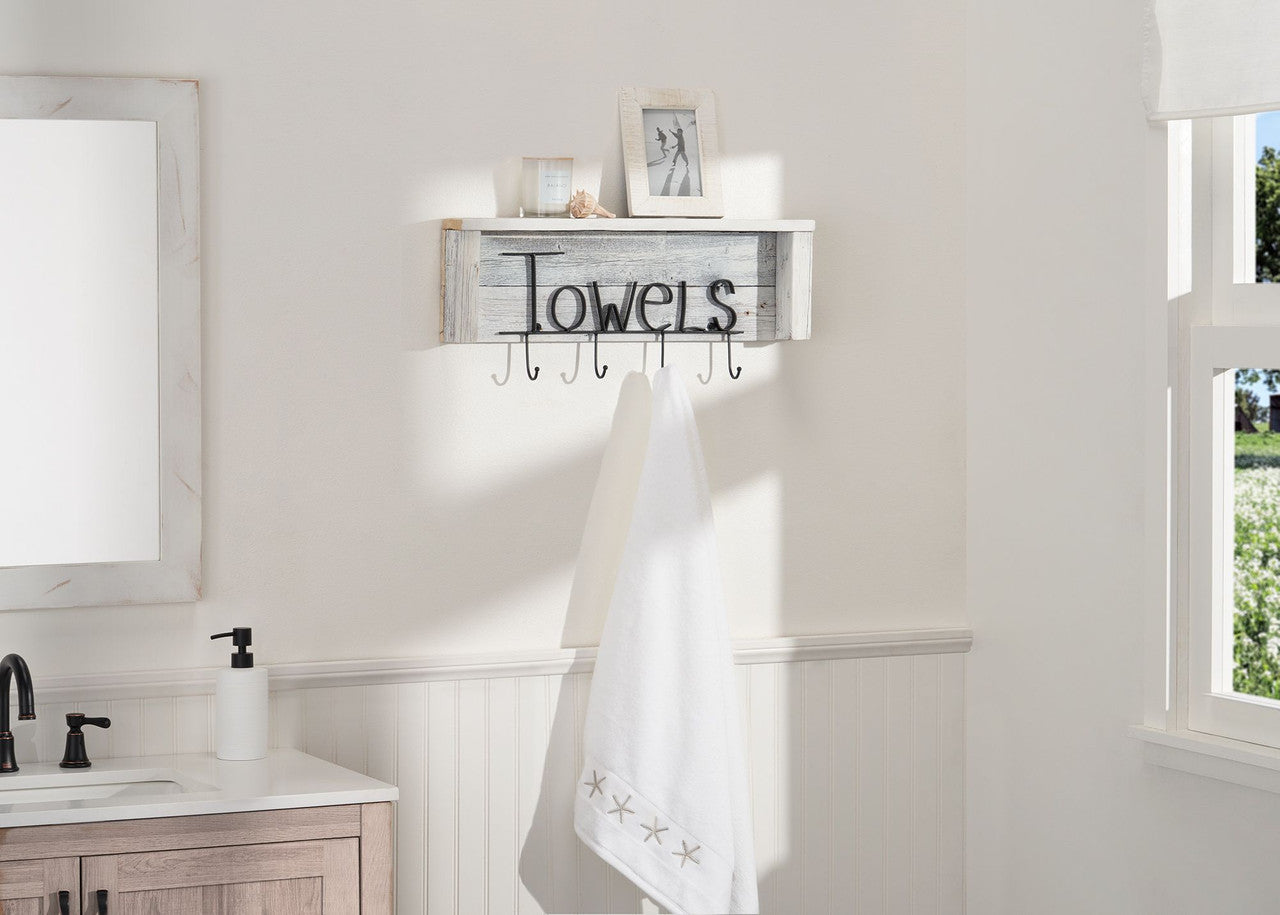 A white wooden shelf with black metal lettering that spells "Towels" is mounted on a white bathroom wall. Below the shelf are five metal hooks, one of which holds a folded white towel with embroidered stars.