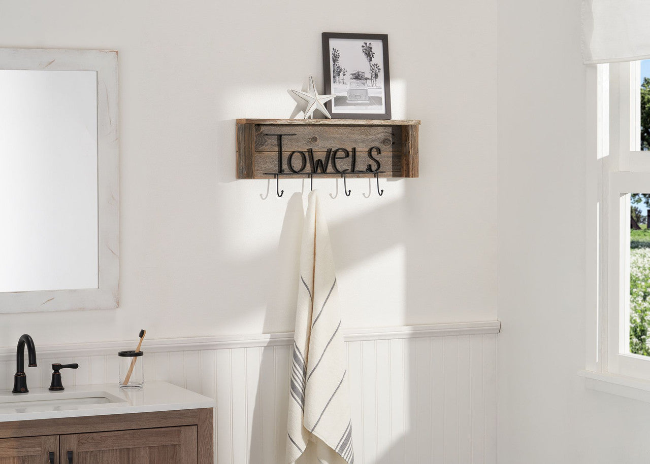 A rustic wooden shelf with the word "Towels" in black script hangs on a white bathroom wall. Below the shelf are six metal hooks, one of which holds a folded white towel with thin blue stripes.