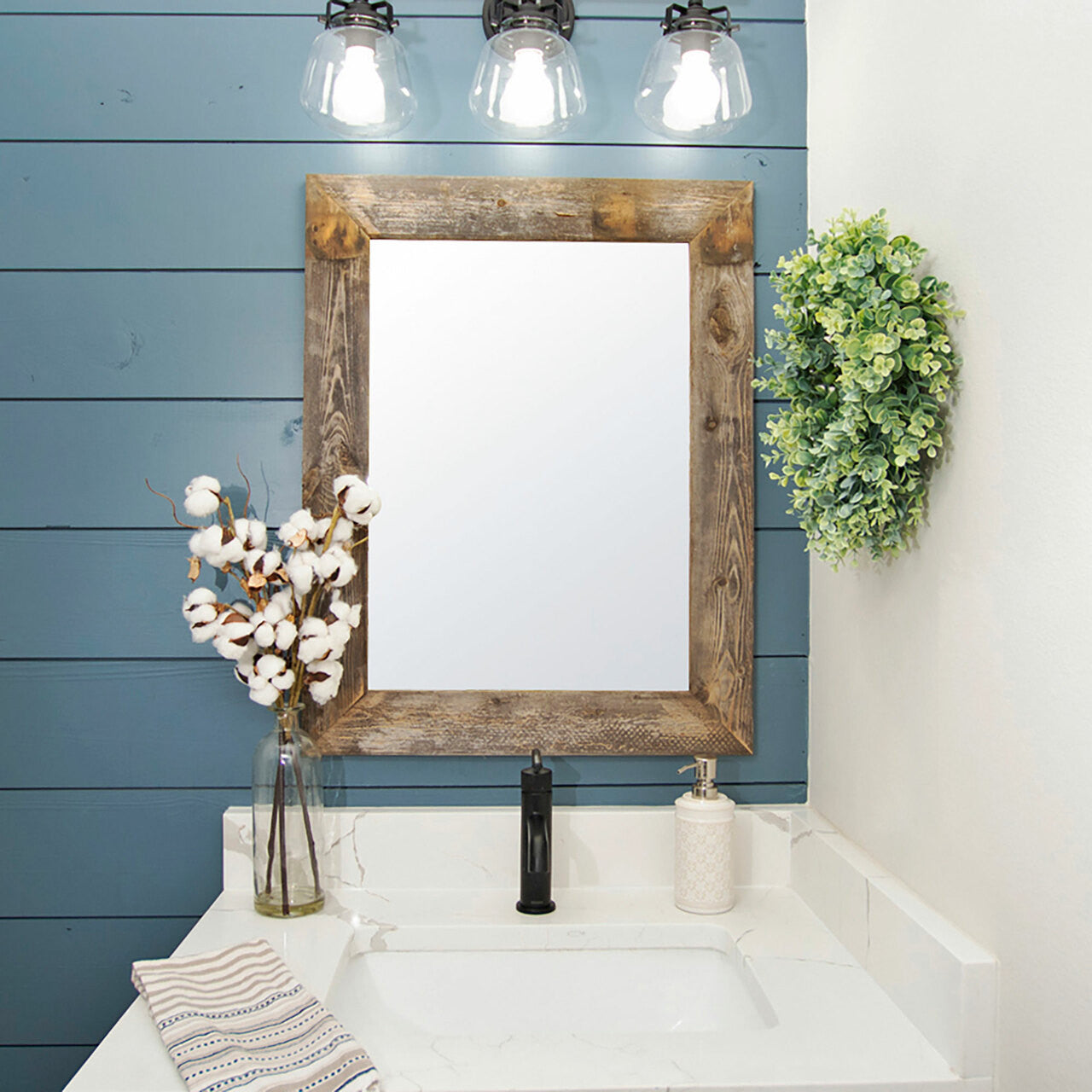 A modern farmhouse style bathroom features a Drakestone barnwood mirror over a white undermount sink with a black faucet. Three industrial-style pendant lights hang above the mirror, and a leafy green wreath and cotton stems add decorative touches.