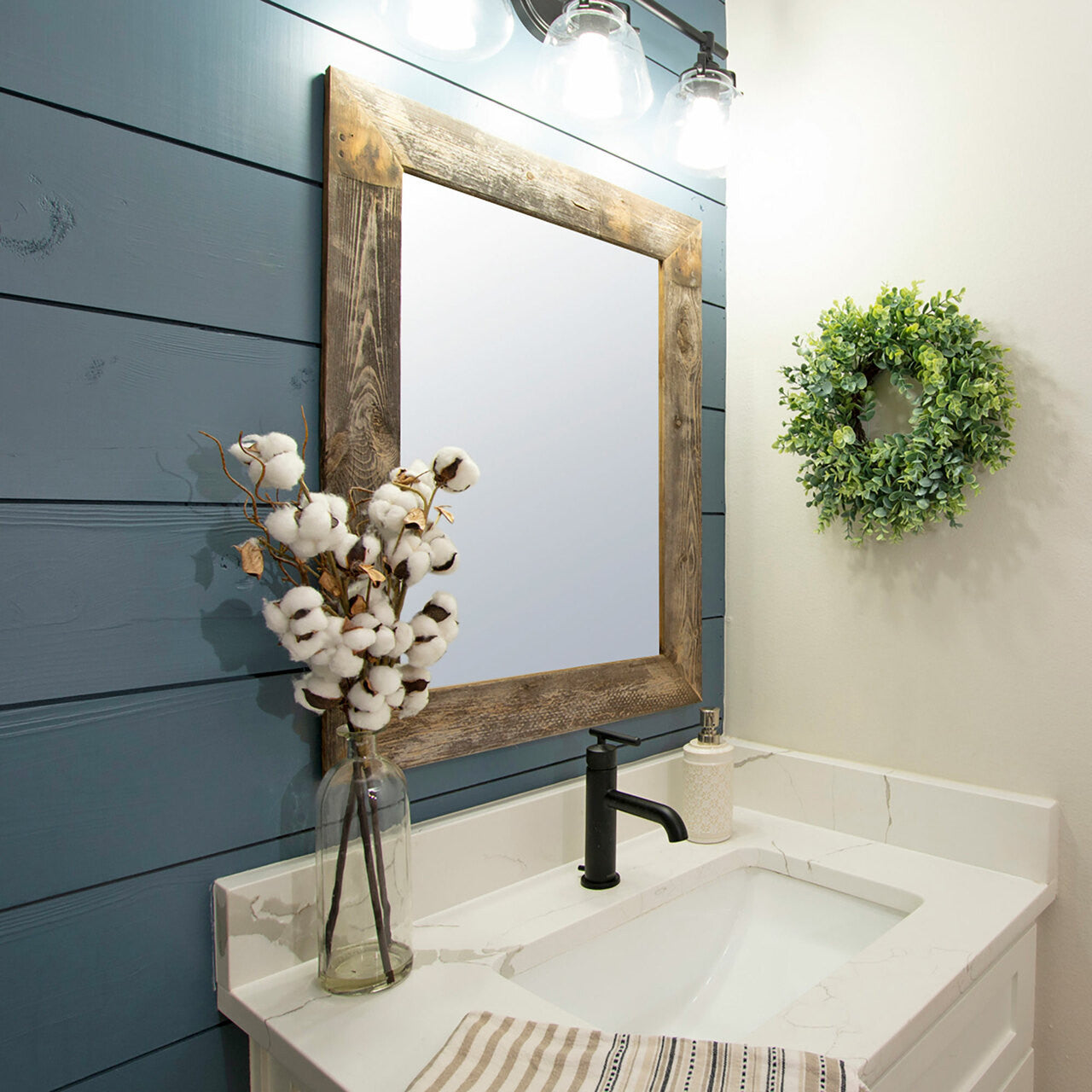 Close-up shot of a bathroom vanity area showcasing a square mirror in a reclaimed wood frame, a white countertop with a black faucet, and a vase of cotton stems against a blue shiplap wall. A small, round green wreath hangs on the adjacent light-colored wall.