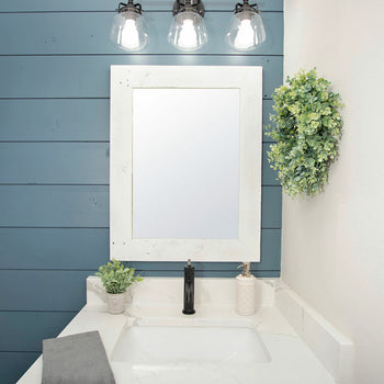 A white-washed wood framed mirror hangs above a bathroom sink with a dark faucet.  The mirror reflects a clear image and is complemented by industrial-style lighting and a decorative green wreath.