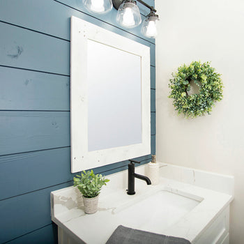 A modern bathroom features a blue shiplap wall, a white framed mirror, and a white vanity with a black faucet. A small green wreath hangs on the adjacent white wall.