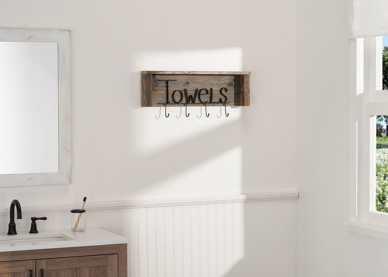 A rustic wooden towel rack with the word "Towels" in black script hangs on a white bathroom wall above a wainscoted lower section. The rack features six metal hooks for hanging towels.