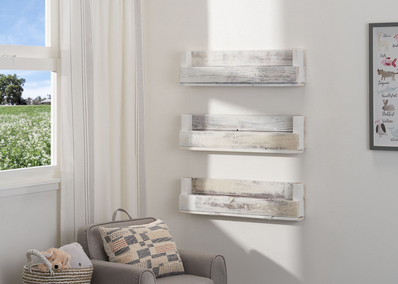 Three weathered, white wooden shelves are mounted on a white wall in a child's room, holding books with colorful spines facing out. A gray armchair with a patterned pillow sits beside a window overlooking a lush green field, with a small woven basket holding stuffed animals in the chair.