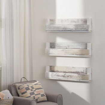 Three weathered, white wooden shelves are mounted on a white wall in a child's room, holding books with colorful spines facing out. A gray armchair with a patterned pillow sits beside a window overlooking a lush green field, with a small woven basket holding stuffed animals in the chair.