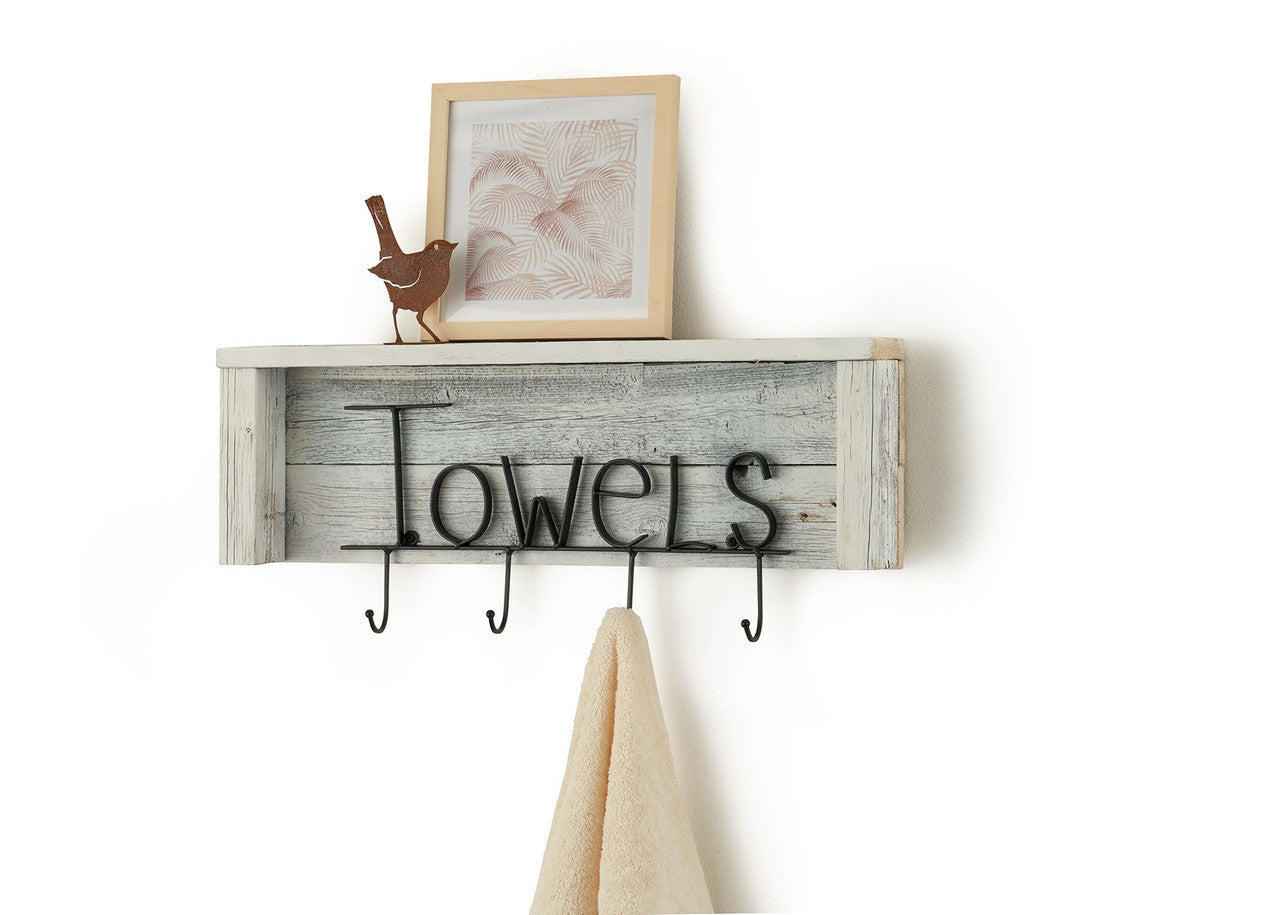 A rustic wooden shelf with a whitewashed plank backing and black metal lettering that spells "Towels" is mounted on a white wall. Below the shelf are four black metal hooks, one of which holds a folded cream-colored towel.