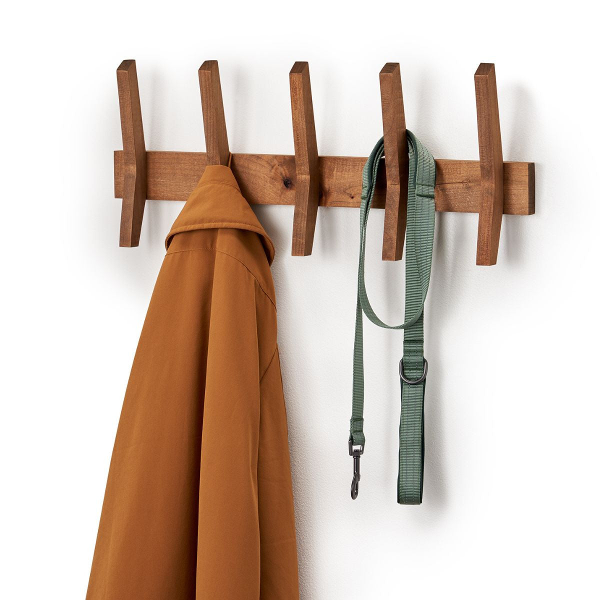 A wooden mid-century coat rack is mounted on a white wall, holding a brown jacket on the leftmost peg and a green dog leash on the fourth peg from the left. The coat rack has five evenly spaced, angled pegs extending from a horizontal bar.