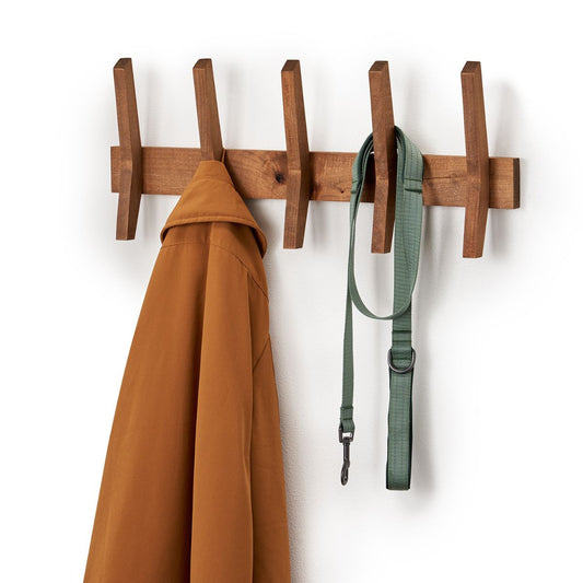 A wooden mid-century coat rack is mounted on a white wall, holding a brown jacket on the leftmost peg and a green dog leash on the fourth peg from the left. The coat rack has five evenly spaced, angled pegs extending from a horizontal bar.