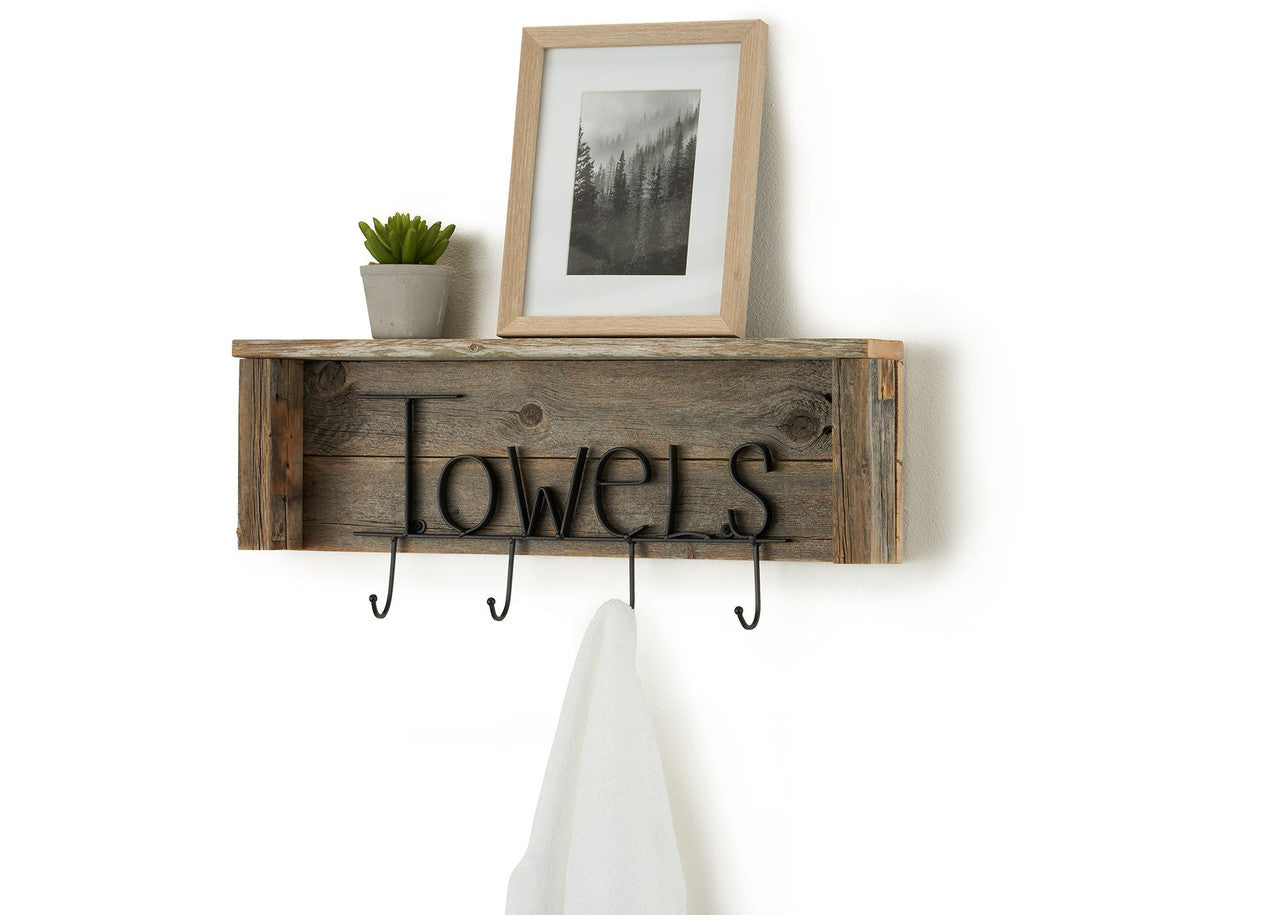 A rustic wooden shelf with the word "Towels" in black metal lettering hangs on a white wall. The shelf has four black metal hooks hanging below it, with a white towel draped over the far right hook.