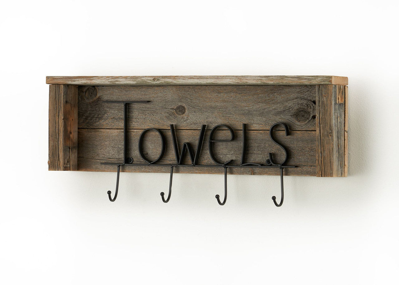 A rustic wooden shelf with a weathered finish has the word "Towels" spelled out in decorative black metal lettering attached to the front. Below the lettering are four black metal hooks for hanging towels.