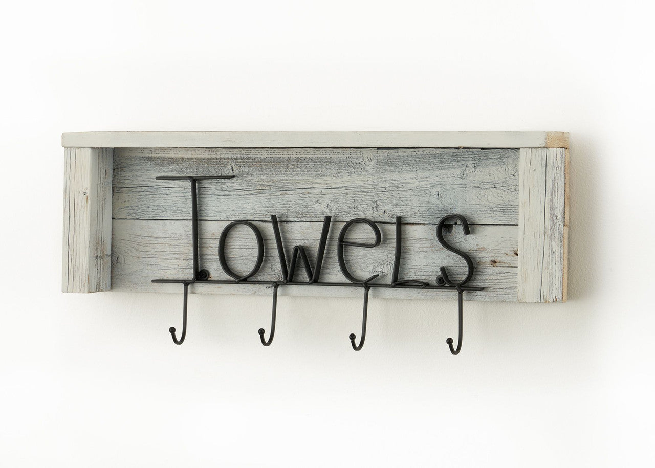 A rustic white wooden shelf with the word "Towels" spelled out in black metal lettering hangs on a white wall. Below the lettering are four black metal hooks for hanging towels.