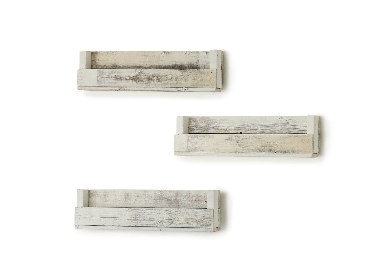 A set of three long, whitewashed wooden shelves are mounted on a white wall, offering a simple storage solution. The shelves have a handmade, rustic look with visible wood grain and slight imperfections.