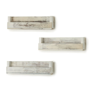 A set of three long, whitewashed wooden shelves are mounted on a white wall, offering a simple storage solution. The shelves have a handmade, rustic look with visible wood grain and slight imperfections.