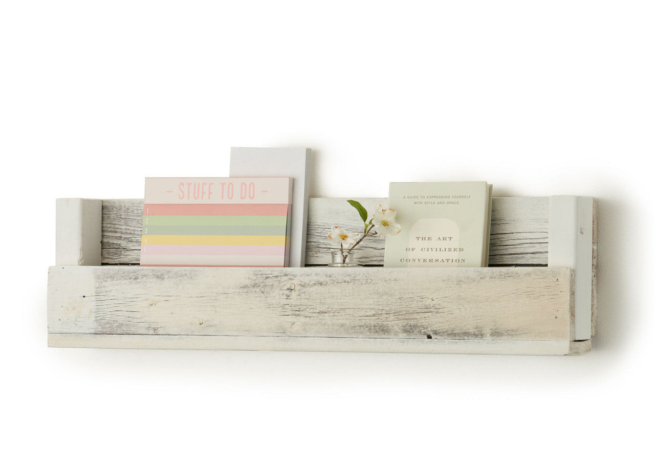 A white wooden shelf with a rustic, weathered finish is mounted on a white wall, holding a notepad titled "STUFF TO DO", a book titled "THE ART OF CIVILIZED CONVERSATION", a small glass vase with a white flower, and a small white notepad. The shelf is made from reclaimed wood and has a simple, minimalist design.