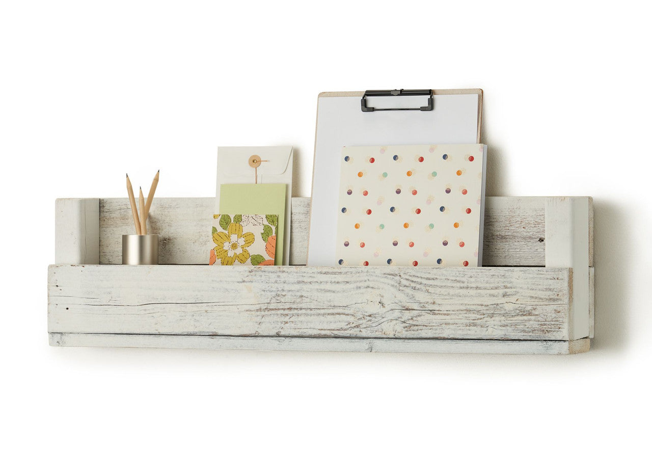 A white, rustic wooden shelf with a back panel is mounted on a white wall. The shelf holds a small metal container with pencils, two greeting cards, a clipboard with a blank white sheet of paper, and a notebook with colorful dots.