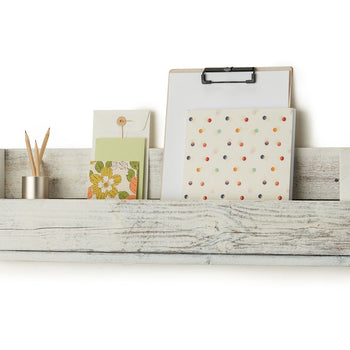 A white, rustic wooden shelf with a back panel is mounted on a white wall. The shelf holds a small metal container with pencils, two greeting cards, a clipboard with a blank white sheet of paper, and a notebook with colorful dots.