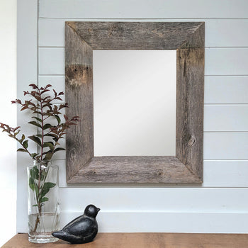 A rustic wooden-framed mirror with a weathered barnwood finish is mounted on a white paneled wall. Below the mirror, a glass vase with leafy branches and a black bird figurine sit on a wooden surface.