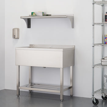 A stainless steel utility sink with a single basin sits on a stand with a lower shelf, positioned against a white wall. To the right, a wire shelving unit holds various kitchen items, and a smaller shelf is mounted on the wall above the sink.