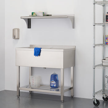 A stainless steel utility sink with a single basin sits on a metal frame, positioned against a white wall. To the right, a metal shelving unit holds various items, and a smaller shelf is mounted on the wall above the sink.