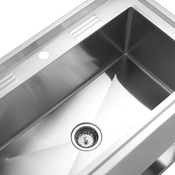 A close-up, angled shot of a stainless steel utility sink basin, showing the drain and a portion of the countertop. The sink is clean and reflective, with a small hole visible on the left side of the countertop.
