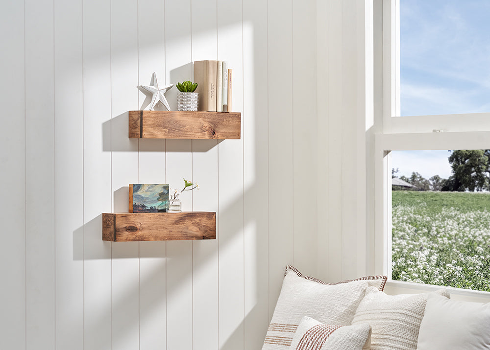 Two thick, floating wooden shelves are mounted on a white shiplap wall next to a bright window overlooking a green field. The shelves are decorated with books, a small potted succulent, and a star ornament, while cozy white pillows rest on a seat below.