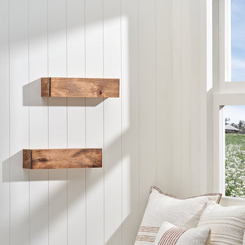 Two floating wooden block shelves are mounted on a white wood-paneled wall, illuminated by sunlight streaming in from an adjacent window. The bright, airy room features neutral-toned decorative pillows in the foreground and a view of a lush green field through the window.