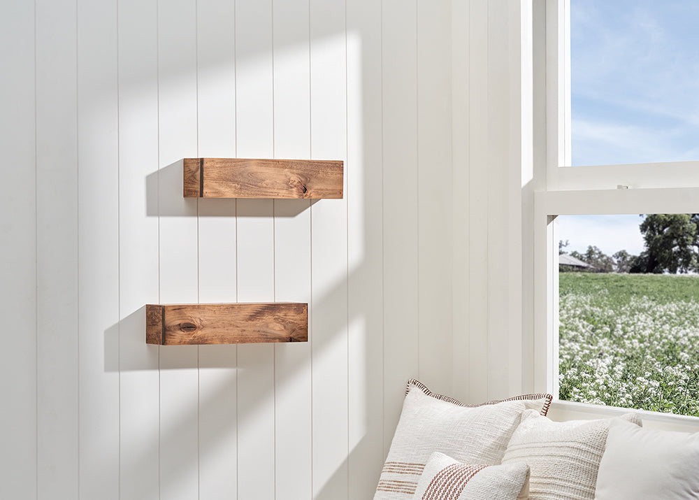 Two floating wooden block shelves are mounted on a white wood-paneled wall, illuminated by sunlight streaming in from an adjacent window. The bright, airy room features neutral-toned decorative pillows in the foreground and a view of a lush green field through the window.