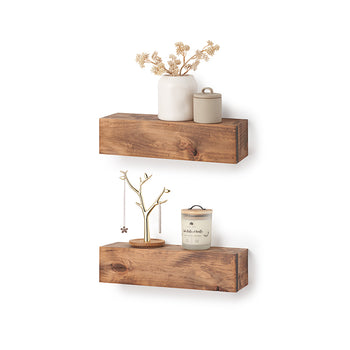 Two thick, rustic wooden floating shelves are mounted on a white wall, displaying various decorative items. The top shelf holds a white vase with dried flowers and a small lidded jar, while the bottom shelf features a gold jewelry tree and a scented candle.
