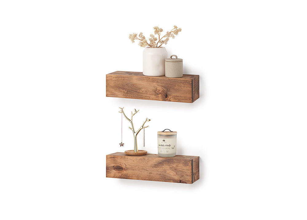 Two thick, rustic wooden floating shelves are mounted on a white wall, displaying various decorative items. The top shelf holds a white vase with dried flowers and a small lidded jar, while the bottom shelf features a gold jewelry tree and a scented candle.