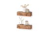 Two thick, rustic wooden floating shelves are mounted on a white wall, displaying various decorative items. The top shelf holds a white vase with dried flowers and a small lidded jar, while the bottom shelf features a gold jewelry tree and a scented candle.