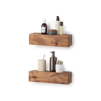 Two floating shelves made of chunky, natural-grained wood are mounted on a white wall and hold various bathroom essentials. The top shelf displays skincare bottles and a small jar, while the bottom shelf holds a toothbrush in a cup, a soap dispenser, and a tiny potted succulent.