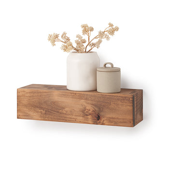 A rectangular, floating wooden shelf with a natural grain and warm brown finish is mounted against a plain white wall. Styled on top of the shelf are a smooth white ceramic vase holding dried, beige floral branches and a small, textured beige canister with a lid.