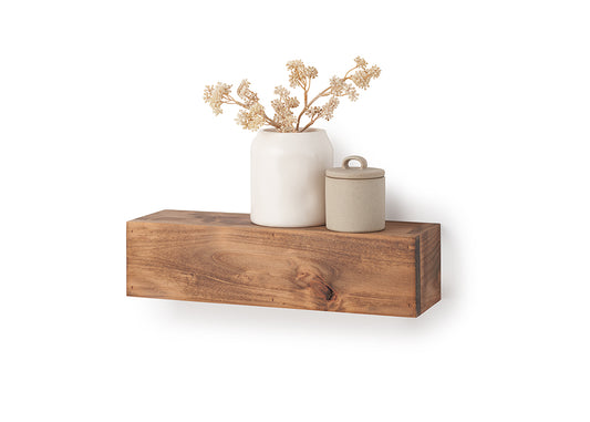 A rectangular, floating wooden shelf with a natural grain and warm brown finish is mounted against a plain white wall. Styled on top of the shelf are a smooth white ceramic vase holding dried, beige floral branches and a small, textured beige canister with a lid.