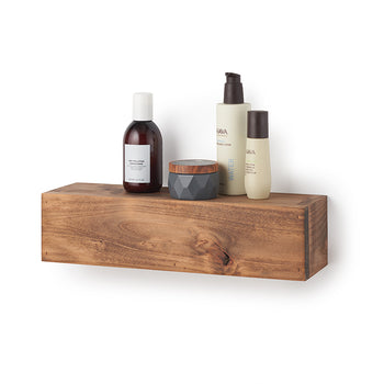A rectangular, floating wooden shelf with a natural grain and warm brown finish is mounted against a plain white wall. Styled on top of the shelf are several skincare items, including a dark amber bottle with a white cap, a small textured grey jar with a wooden lid, and two tan-colored 