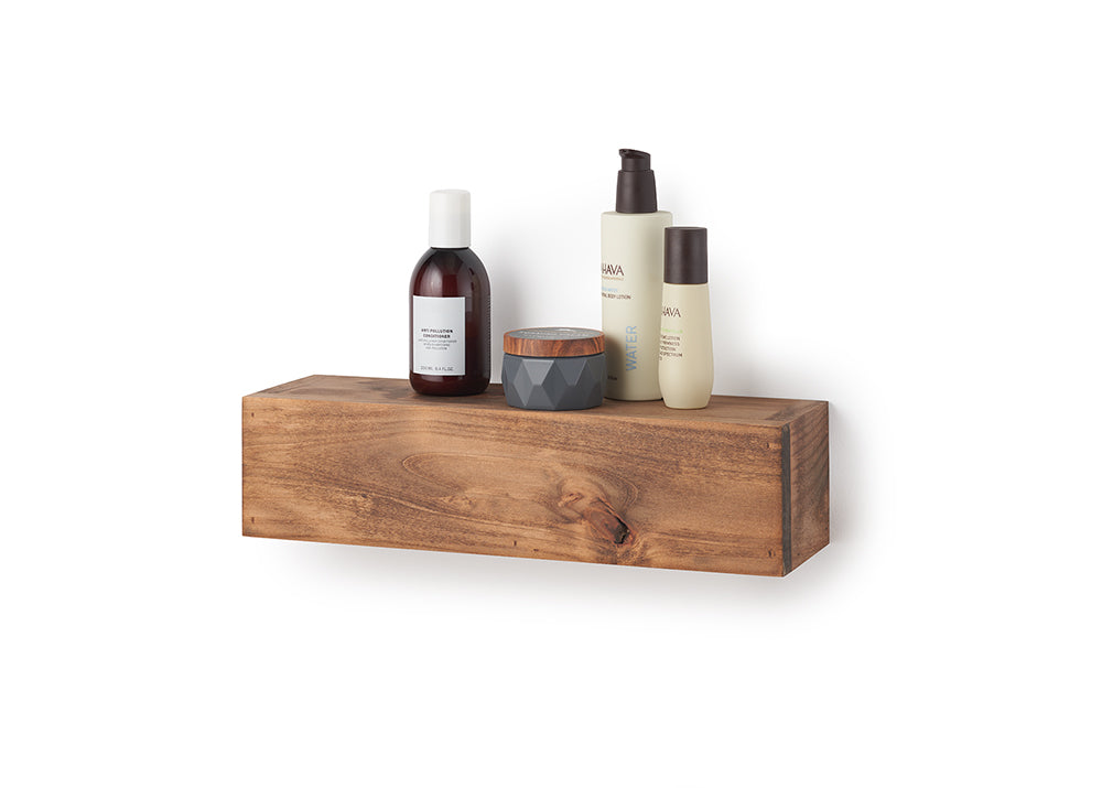 A rectangular, floating wooden shelf with a natural grain and warm brown finish is mounted against a plain white wall. Styled on top of the shelf are several skincare items, including a dark amber bottle with a white cap, a small textured grey jar with a wooden lid, and two tan-colored "AHAVA" bottles.