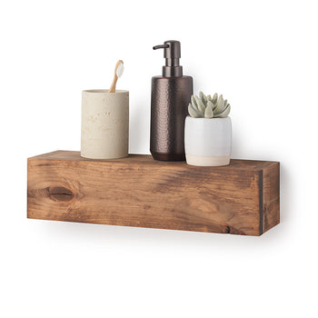 A rectangular, floating wooden shelf with a natural grain and warm brown finish is mounted against a plain white wall. Styled on top of the shelf are bathroom accessories, including a beige textured tumbler holding a wooden toothbrush, a dark metallic soap dispenser, and a small faux succulent in a white and tan ceramic pot.