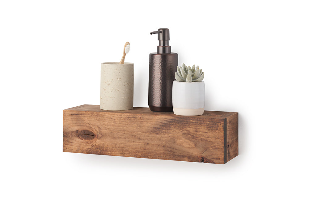 A rectangular, floating wooden shelf with a natural grain and warm brown finish is mounted against a plain white wall. Styled on top of the shelf are bathroom accessories, including a beige textured tumbler holding a wooden toothbrush, a dark metallic soap dispenser, and a small faux succulent in a white and tan ceramic pot.
