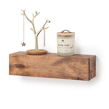 A rectangular, floating wooden shelf with a natural grain and warm brown finish is mounted against a plain white wall. Styled on top of the shelf are a gold-finished, tree-shaped jewelry stand on a round wooden base and a frosted glass jar candle labeled 
