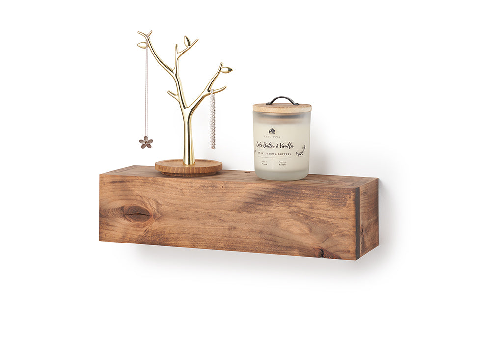 A rectangular, floating wooden shelf with a natural grain and warm brown finish is mounted against a plain white wall. Styled on top of the shelf are a gold-finished, tree-shaped jewelry stand on a round wooden base and a frosted glass jar candle labeled "Cake Batter & Vanilla" with a light wooden lid.