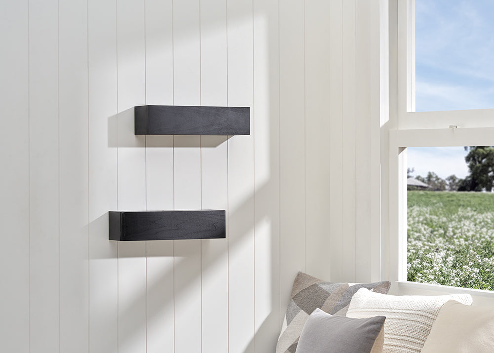 Two matte black floating shelves are mounted vertically on a white shiplap wall next to a bright window. The scene is softly lit, featuring neutral-toned decorative pillows in the foreground and a view of a green field through the window.