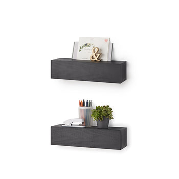Two wall-mounted, charcoal-colored floating shelves are stacked vertically against a plain white background. The top shelf displays a card and a gold ampersand decor piece, while the bottom shelf holds a stapler, a cup of pens, and a small potted succulent.