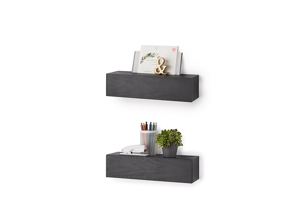 Two wall-mounted, charcoal-colored floating shelves are stacked vertically against a plain white background. The top shelf displays a card and a gold ampersand decor piece, while the bottom shelf holds a stapler, a cup of pens, and a small potted succulent.