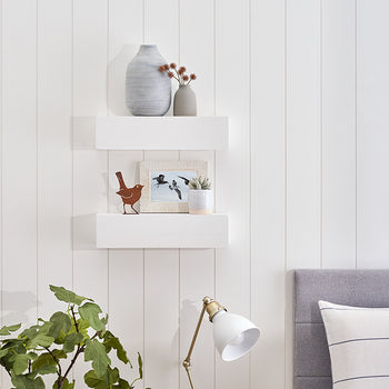 Two white floating shelves are mounted on a white wood-paneled wall, decorated with vases, a small bird figurine, and a framed photo. Below the shelves, a green leafy plant and a white task lamp sit near a gray upholstered headboard with a striped pillow.