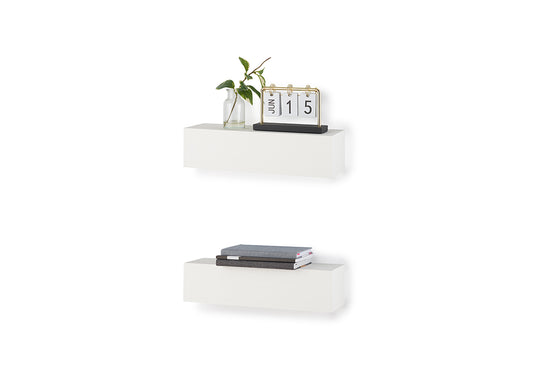 Two minimalist white floating block shelves are mounted vertically on a plain white wall, holding decorative items. The top shelf displays a small glass bud vase with green leaves and a flip-style desk calendar, while the bottom shelf holds a stack of three books.