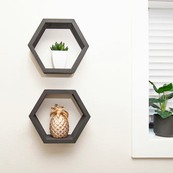 Two hexagonal black wall shelves are mounted on a white wall, one above the other. The top shelf holds a small white pot with a green succulent, while the bottom shelf displays a gold decorative pineapple.
