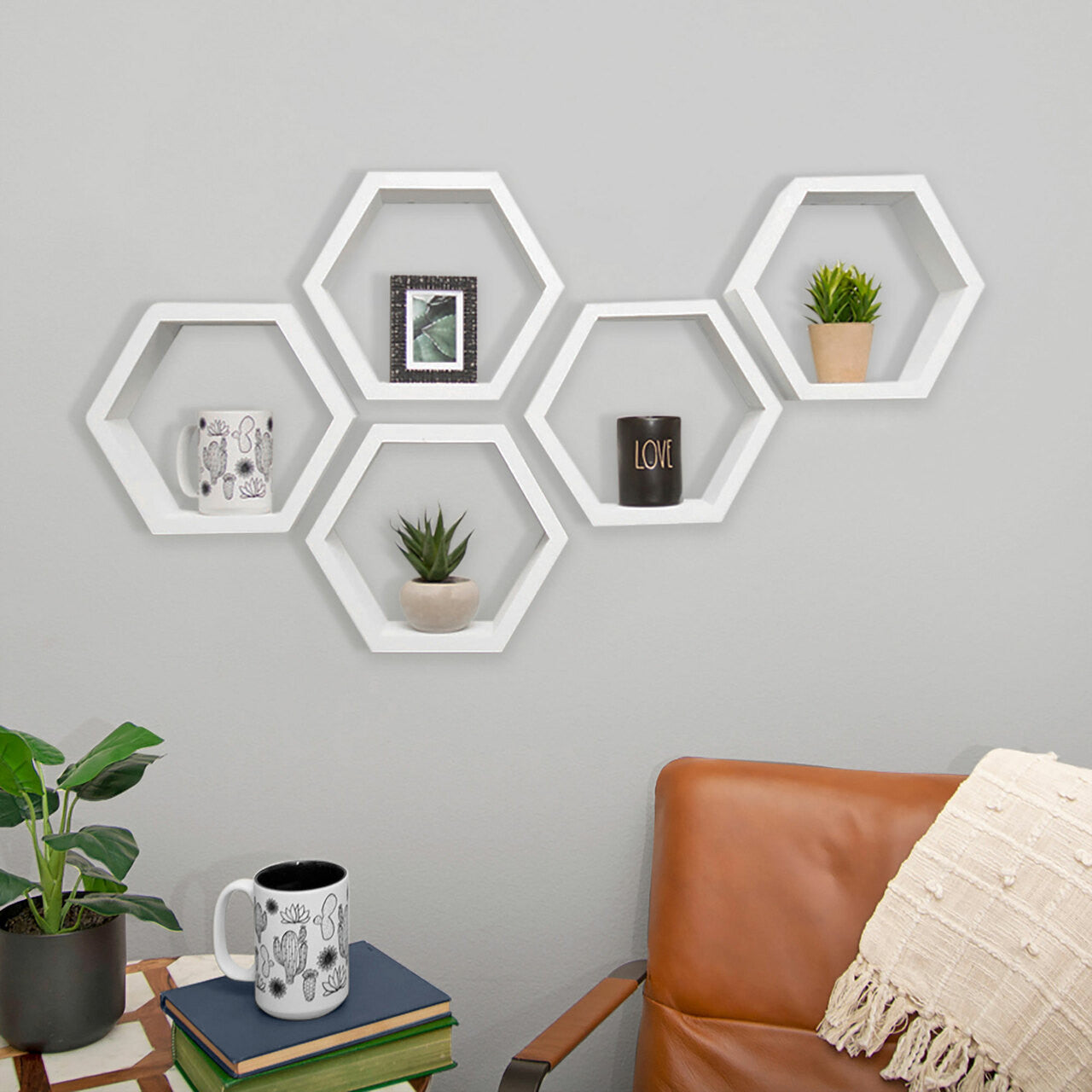 Five white hexagon shelves arranged asymmetrically on a light gray wall.  They hold a small framed picture, a potted plant, a mug, another plant, and a small black container labeled "LOVE".