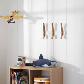 A children's room corner features a wooden cube shelf unit with books, a patterned cylindrical container, and a plush blue shark toy on top. Above the shelf, a toy yellow biplane hangs from the white paneled wall, which also displays a modern wooden and white coat rack and a framed space-themed picture.