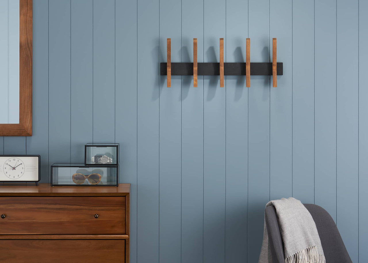 A modern wall-mounted coat rack with wooden pegs is centered on a blue paneled wall. A dresser, mirror, clock, and chair are visible in the surrounding room.